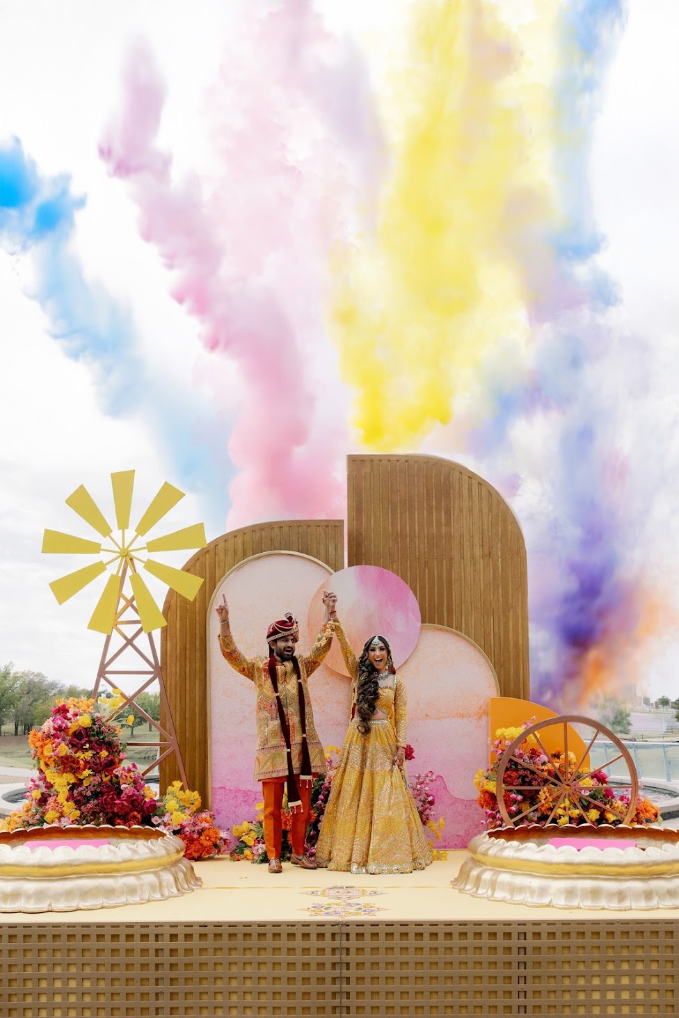Two people in traditional attire stand on a decorated stage with colorful smoke and floral arrangements.