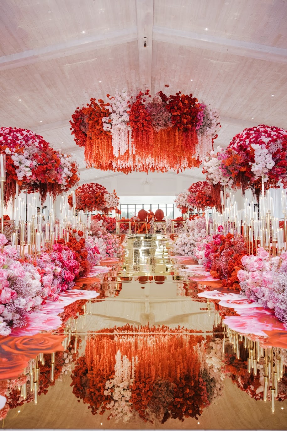 Elegant event space decorated with pink and red flowers, floral arrangements, and a reflective gold floor, creating a symmetrical scene.