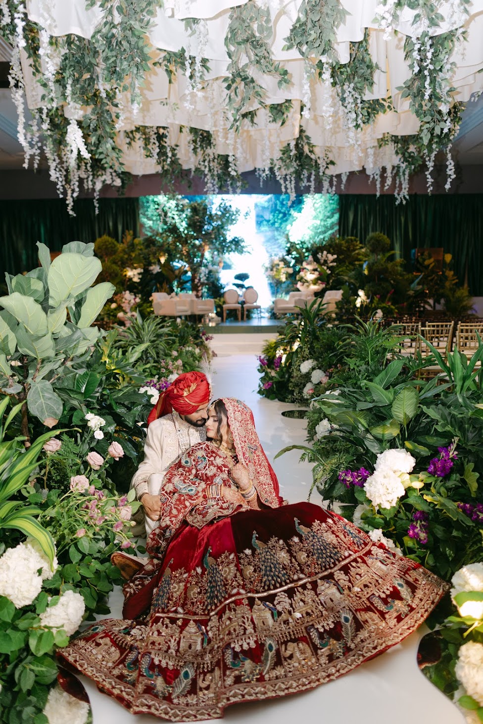 Person in traditional attire sitting among lush floral decorations with a bright outdoor scene in the background.