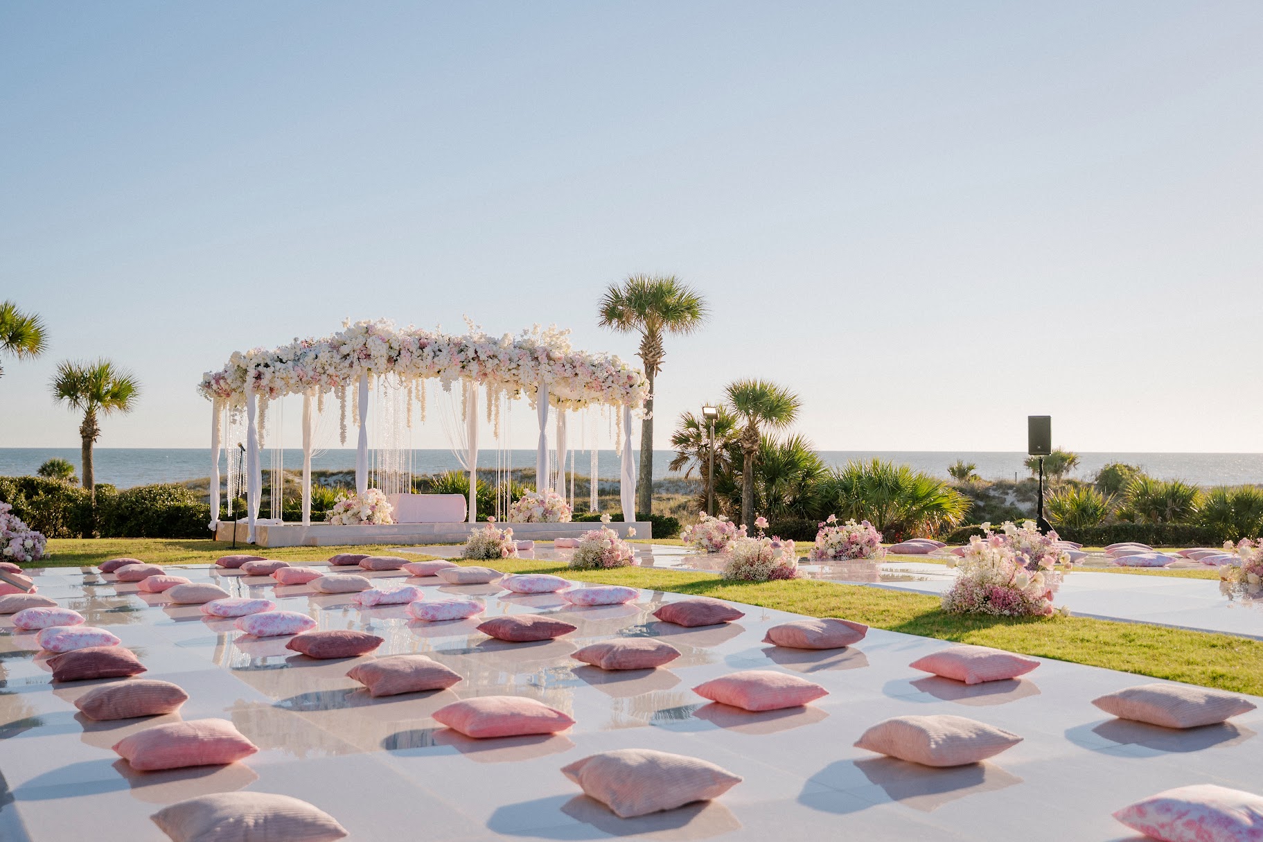Outdoor wedding setup with a decorated canopy, pink cushions on the ground, and palm trees near the ocean.