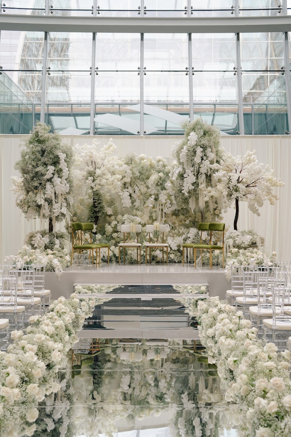 Indoor wedding setup with white floral arrangements, a reflective aisle, and a glass ceiling, in a modern building.
