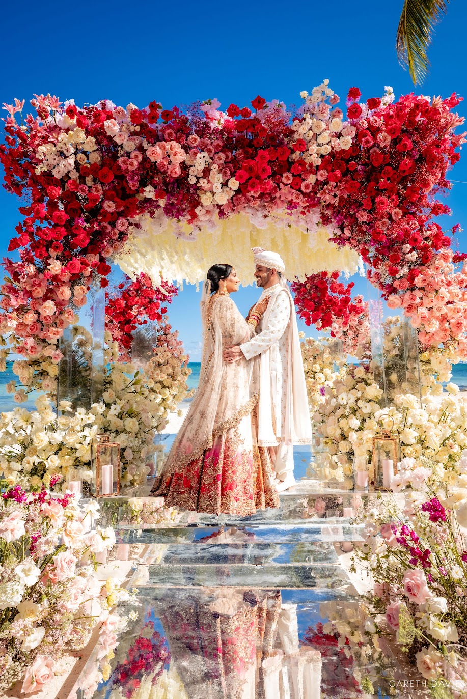 Couple in traditional attire standing under a floral arch at a wedding ceremony, surrounded by colorful flowers and a blue sky.