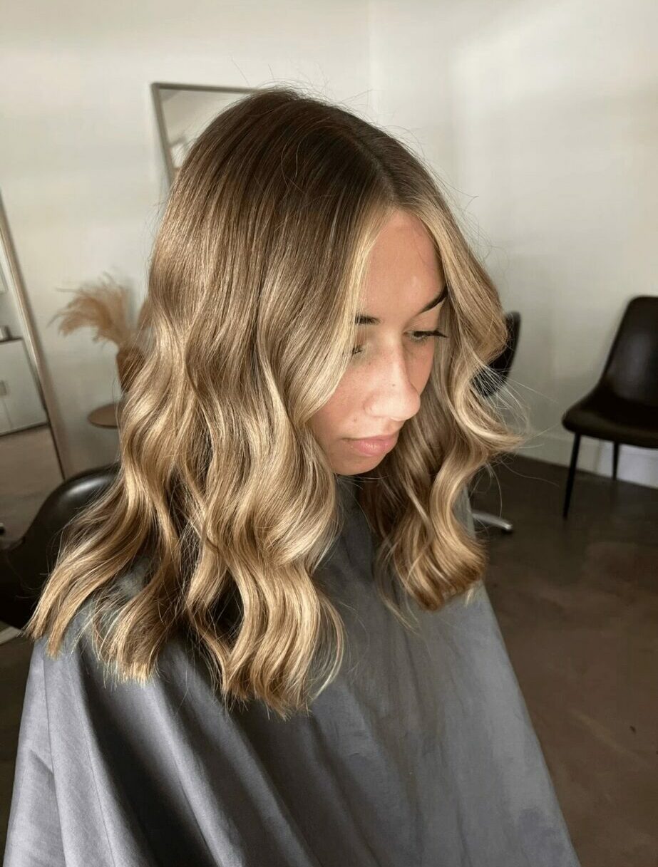 Woman with wavy blonde hair looking down, sitting in a salon chair, with a mirror and chair in background.