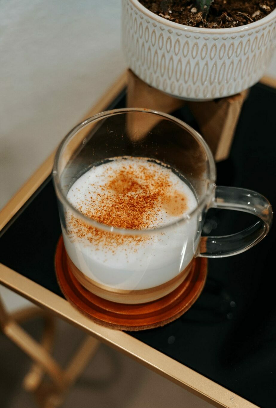 Clear glass mug with frothy beverage topped with cinnamon, on a wooden tray with a potted plant nearby.