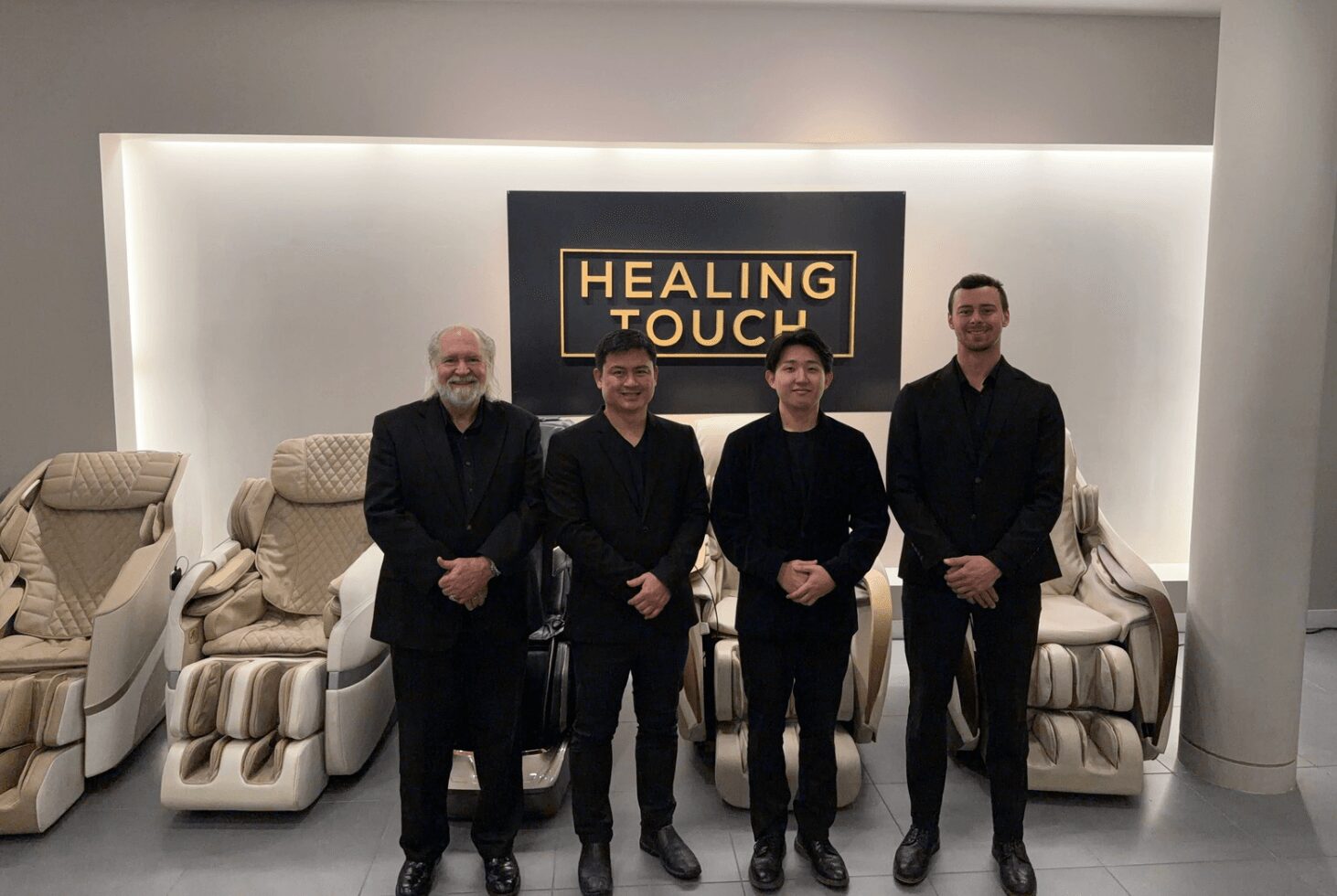 Four people stand in front of a sign that reads 'Healing Touch' in a modern, well-lit room with massage chairs.