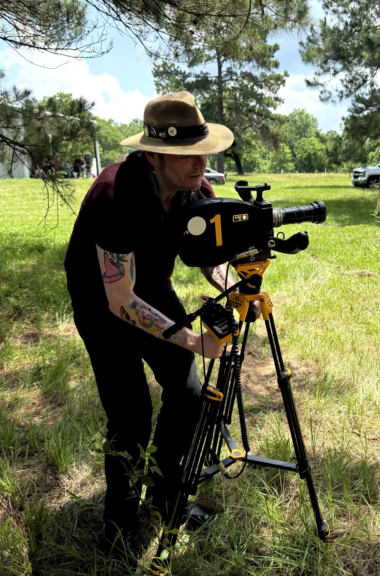 Person wearing a hat operating a camera on a tripod outdoors with trees and grass.