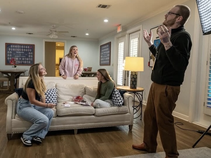 Four people in a living room, one standing and three sitting on a sofa, with a lamp and windows in the background.