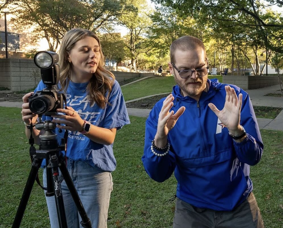 Two people outdoors, one woman adjusting camera on tripod, one man gesturing with hands, trees and park in background.