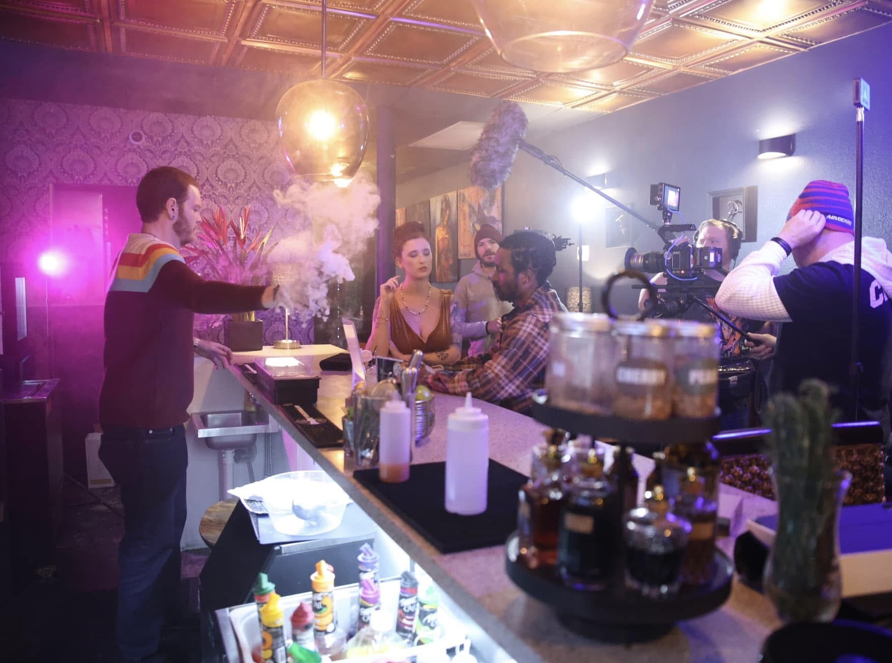 People in a bar or club with colorful lighting, some sitting at the counter, others standing, and a person with a rainbow sweater.