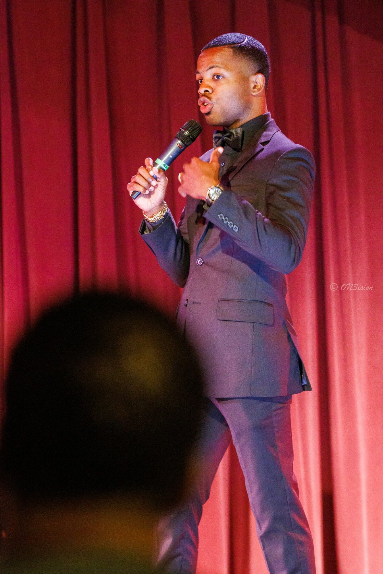 Young man in a suit holding a microphone on stage with red curtains behind him.