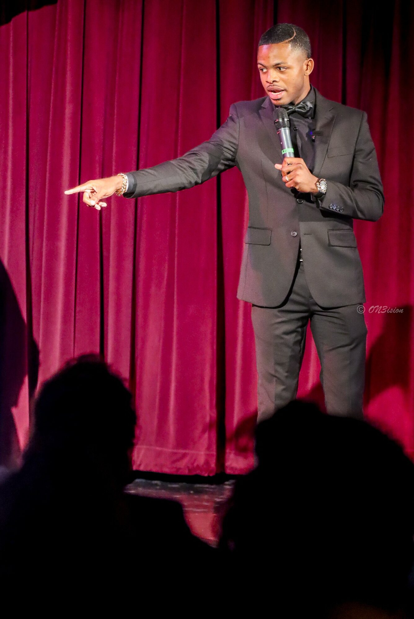 Male comedian in a suit holding a microphone, pointing to the left on stage with red curtains behind him.
