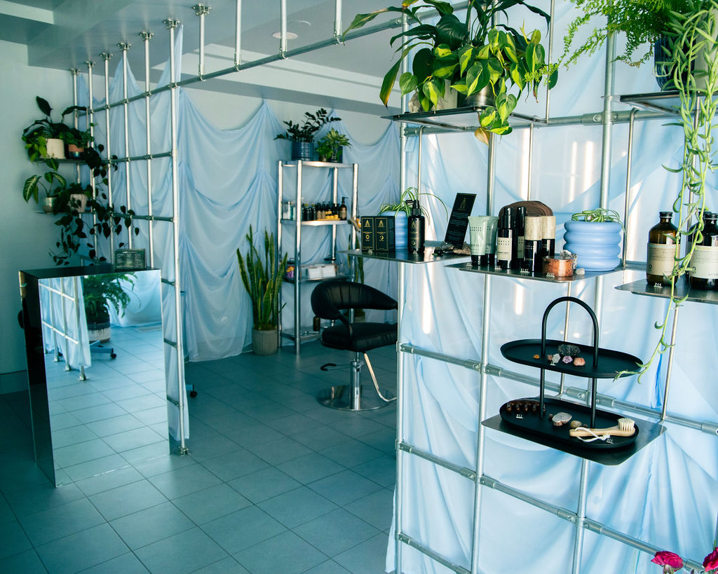 Indoor space with shelves, plants, and chairs, decorated with white fabric and greenery, sunlight streaming in.