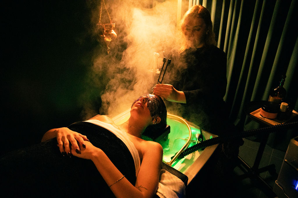 Person lying down with eyes closed, receiving a facial treatment in a dimly lit room with steam and a therapist holding tools.
