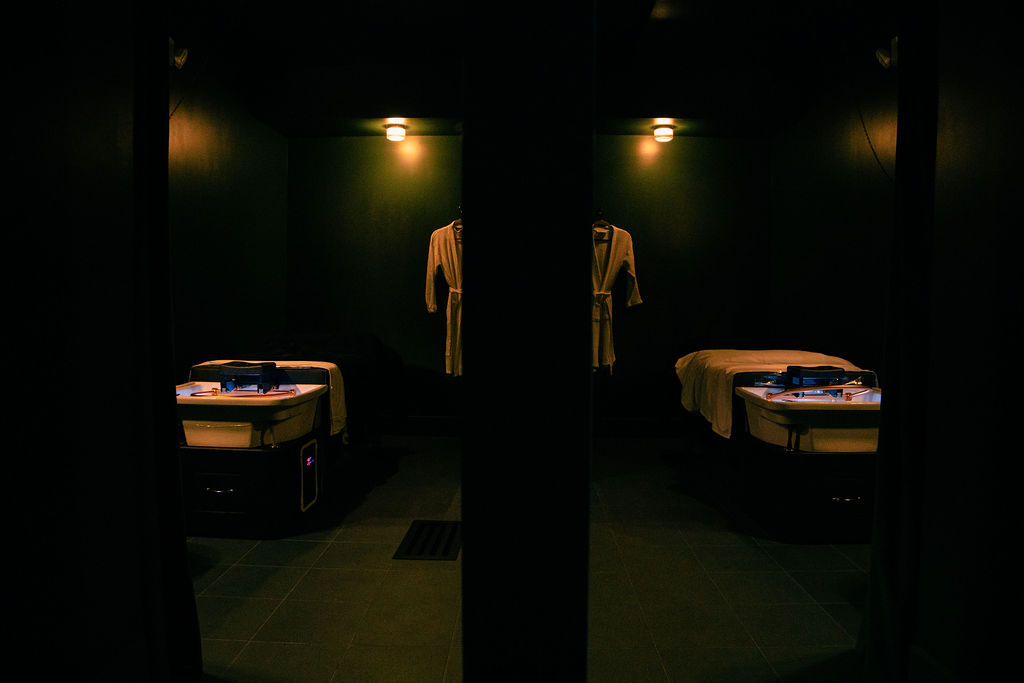 Two hospital beds in a dimly lit room with a mirror reflecting a shirt hanging on the wall.