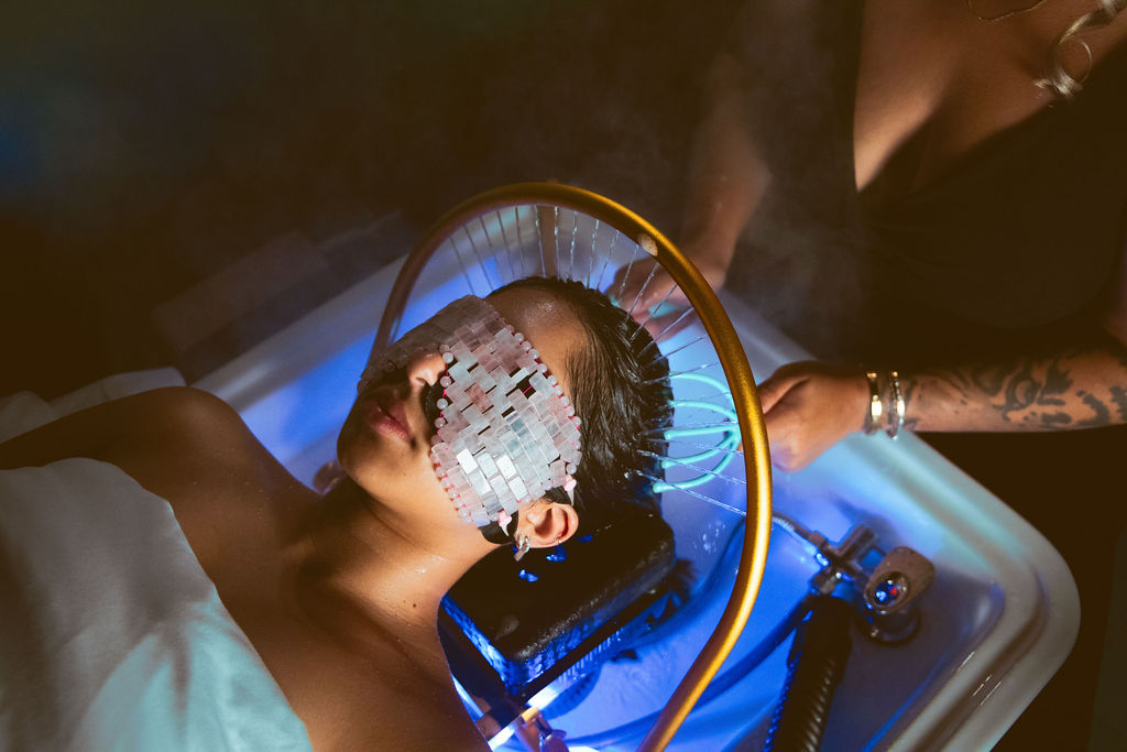Person with mosaic eye covering lying in a sink with blue lighting, another person standing nearby holding a tool.