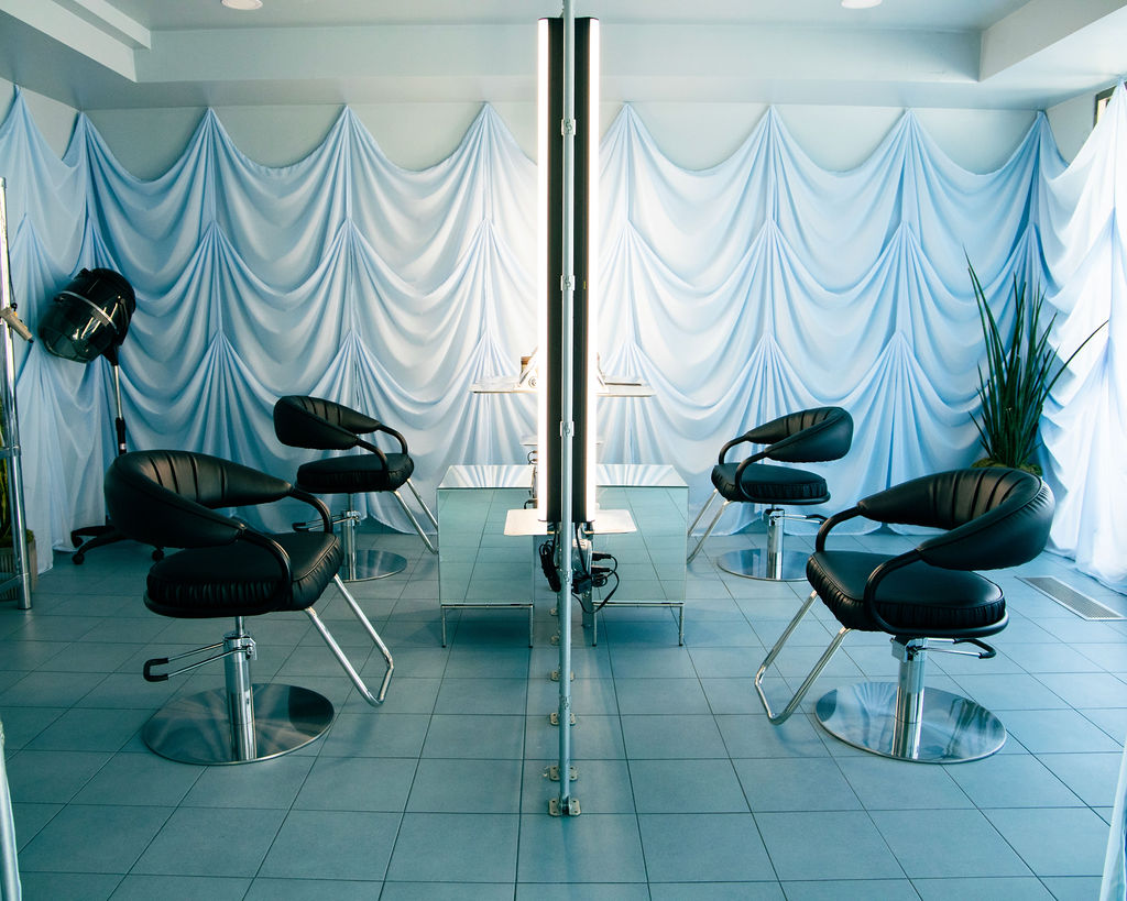 Salon with four black chairs, blue draped curtains, a potted plant, and a mirror dividing the space.
