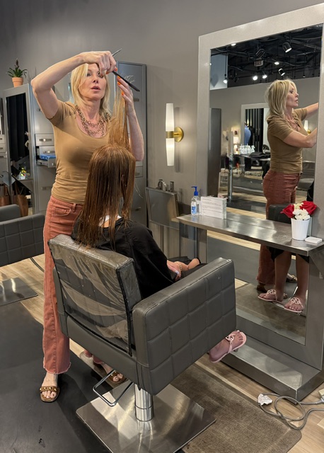 Woman with blonde hair styles a person's hair in a salon, with a mirror reflecting her and the client, and a small flowerpot on the counter.