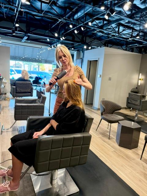 Hairdresser with blonde hair styling a woman's long hair in a salon with modern furniture and large windows.