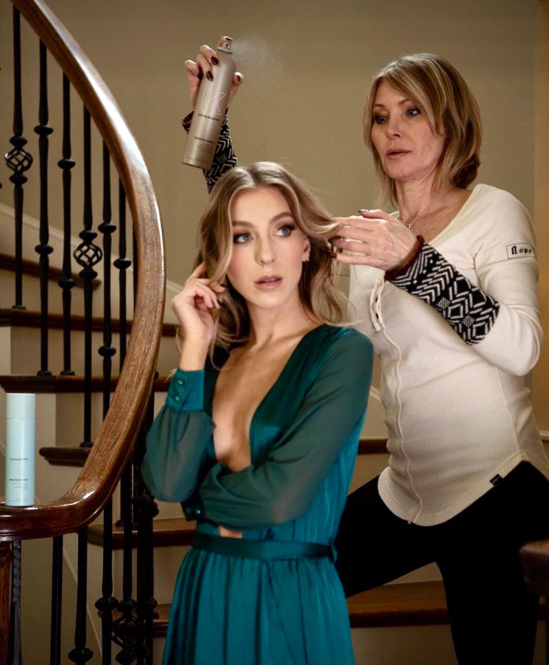 A woman styles a young woman's hair on a staircase landing, with a wooden staircase railing visible on the left.