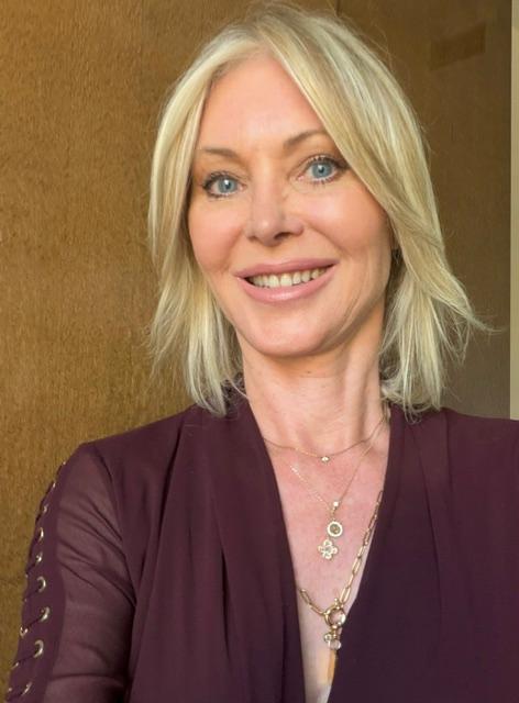 Woman with blonde hair smiling, wearing a dark top and layered necklaces, standing indoors with a brown background.