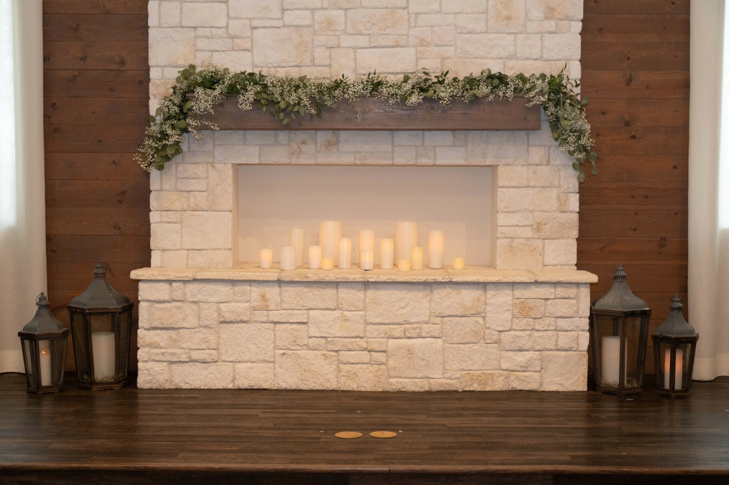 Stone fireplace with candles inside, decorated with greenery and flanked by lanterns on a wooden floor.