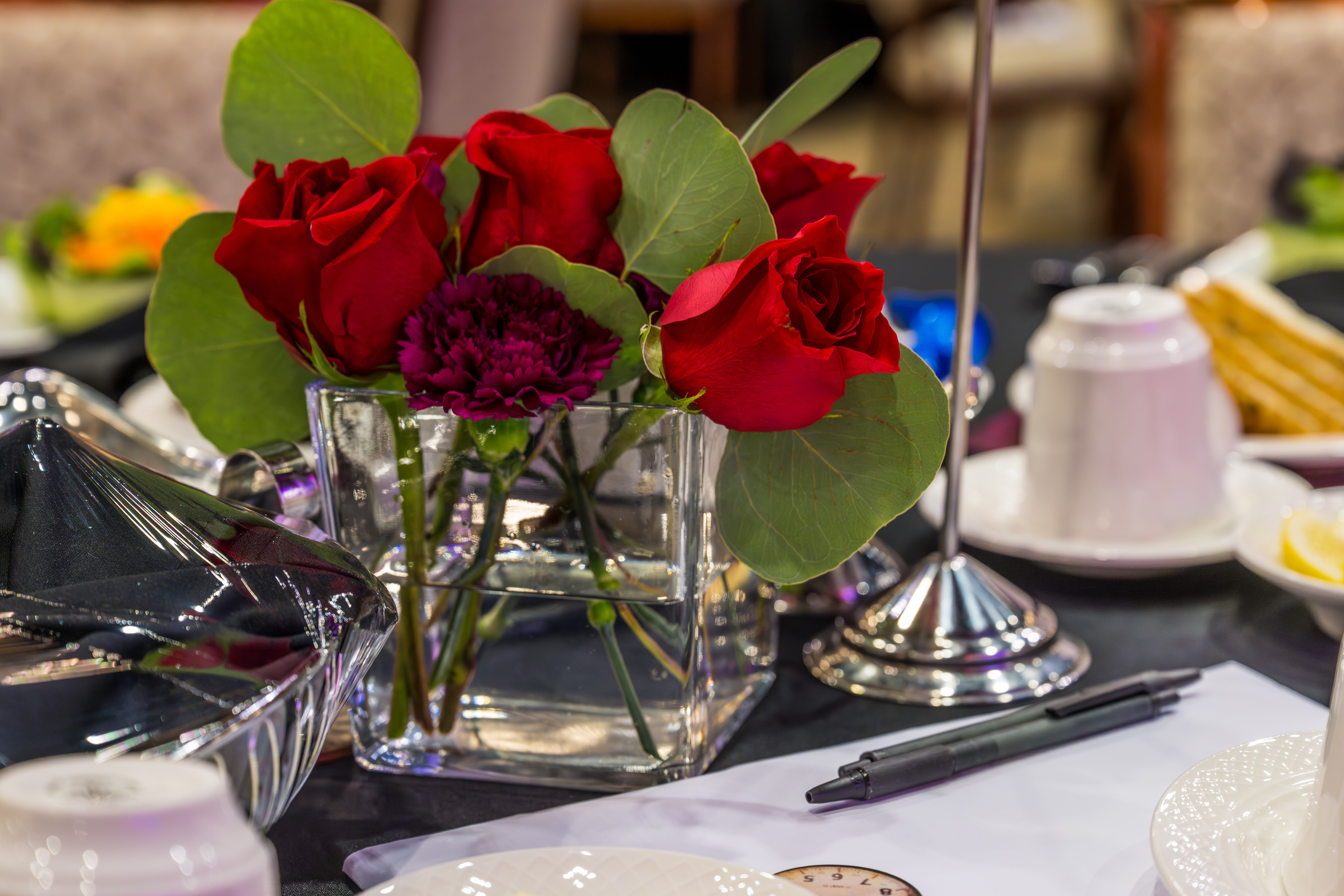 Red roses and purple flowers in a glass vase on a table with various dishes and utensils