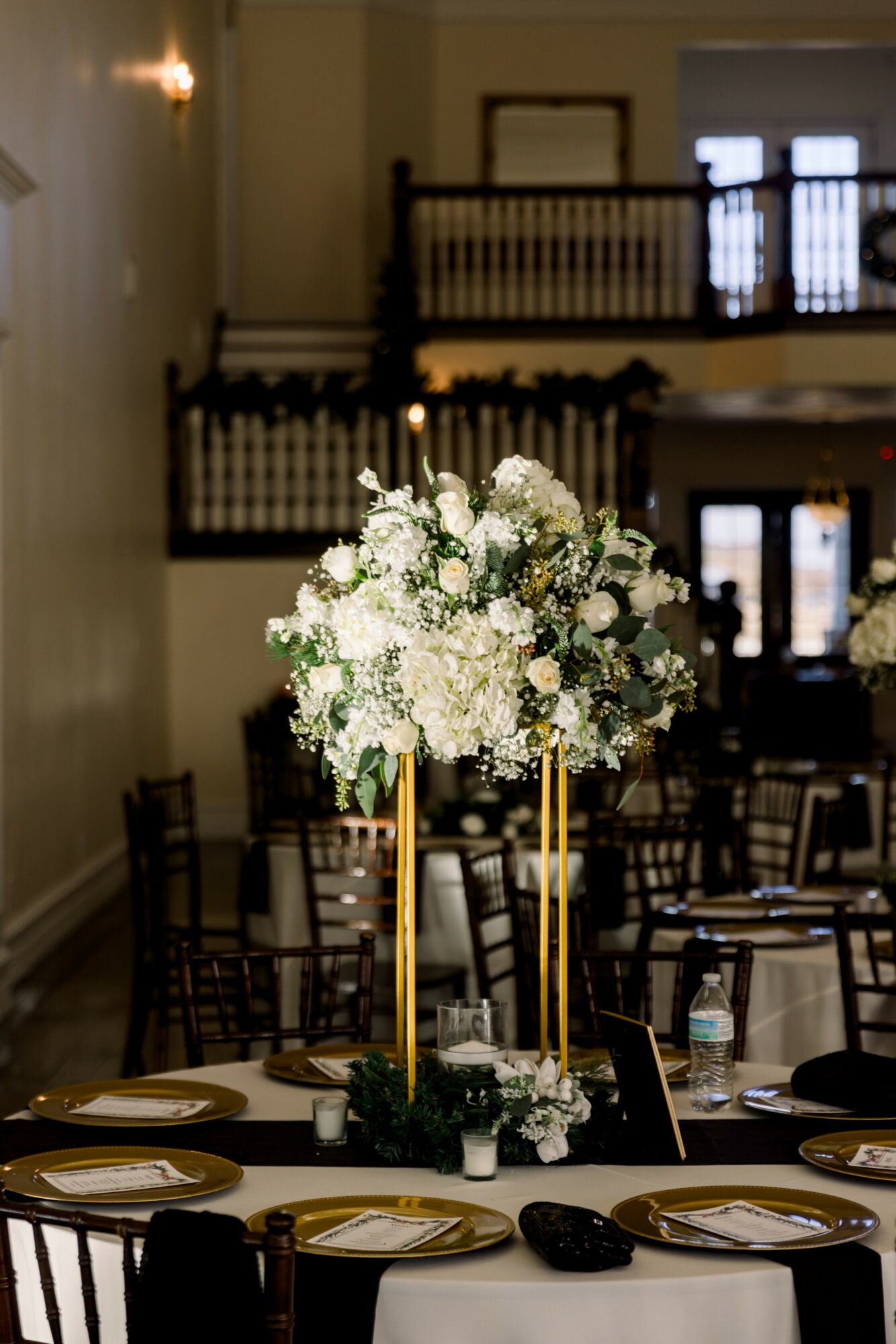 Elegant table setting with a large floral centerpiece and tall gold candleholders in a spacious event hall.