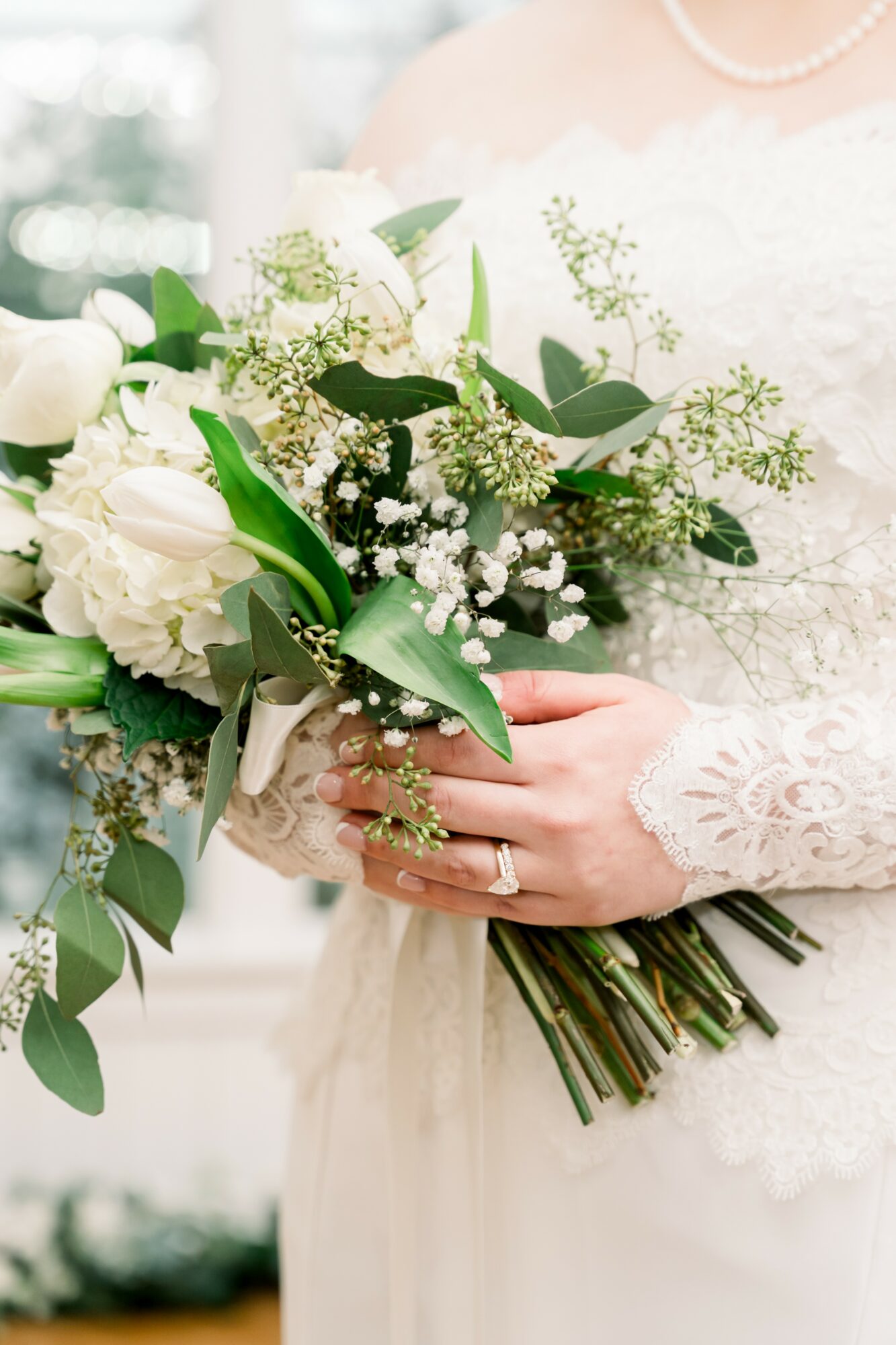 Person holding a bouquet of white flowers and greenery, wearing a lace dress and pearl necklace.