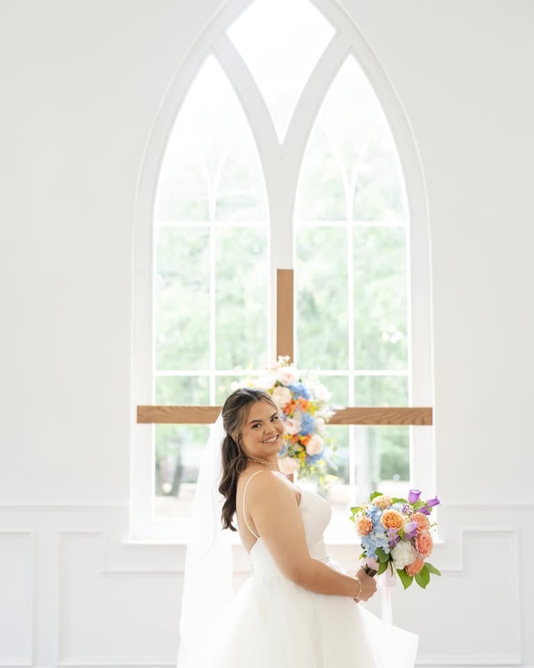 Woman in white dress holding a bouquet in front of a large arched window with sunlight streaming in.