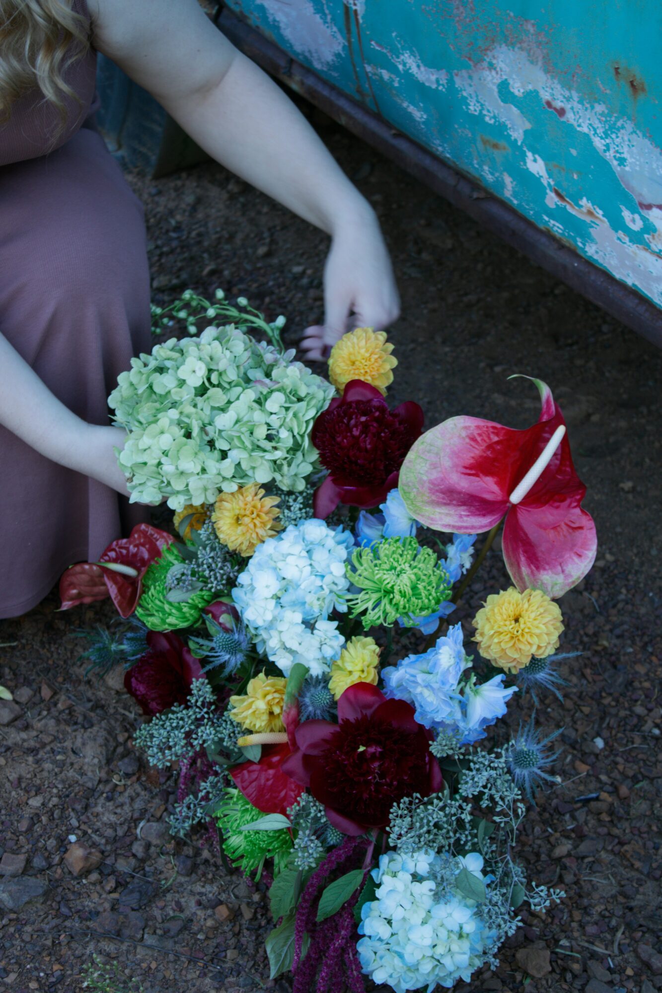 Person arranging colorful flowers on ground near a weathered blue surface, wearing a purple dress, with only hands visible.