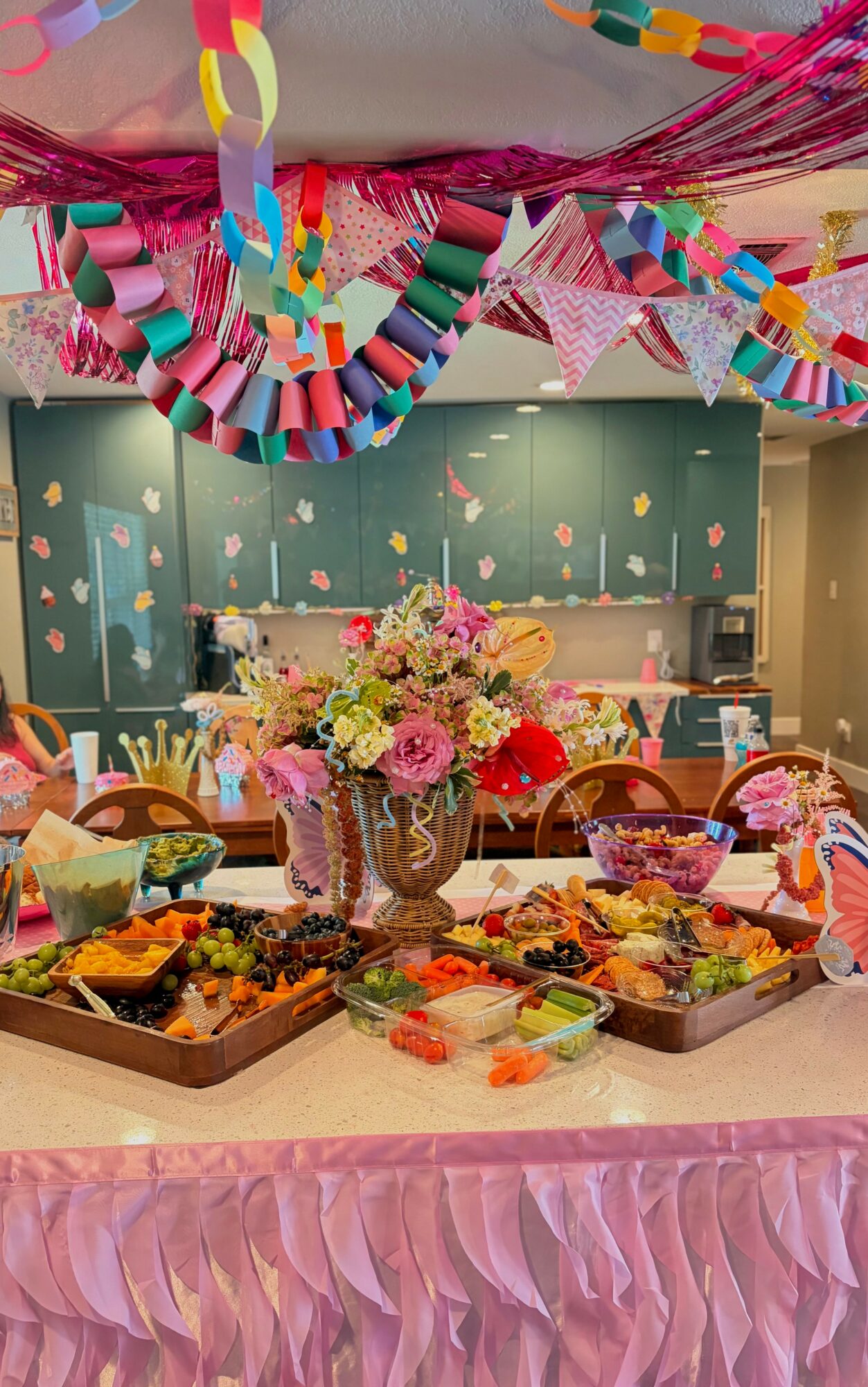 Party table with a floral centerpiece, food trays, and colorful decorations hanging from the ceiling, in a decorated room.