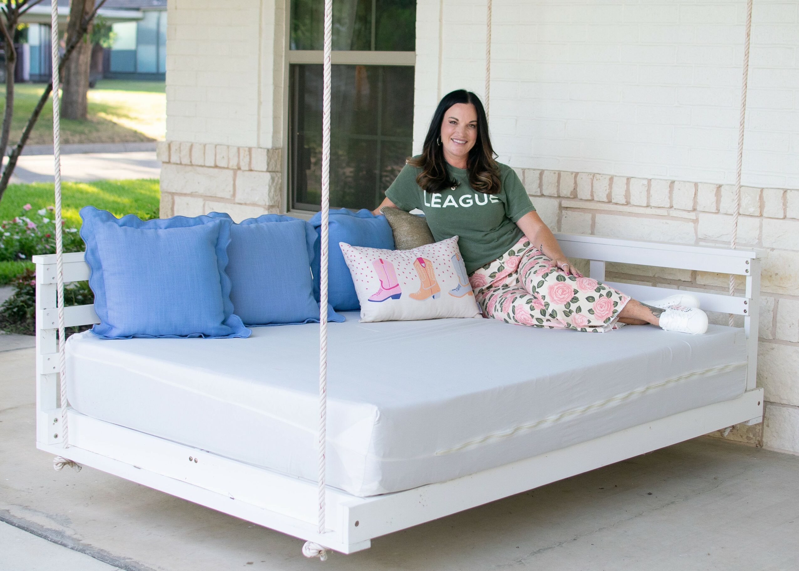 Woman sitting on a white outdoor bed with blue pillows and a decorative pillow, in front of a house wall.