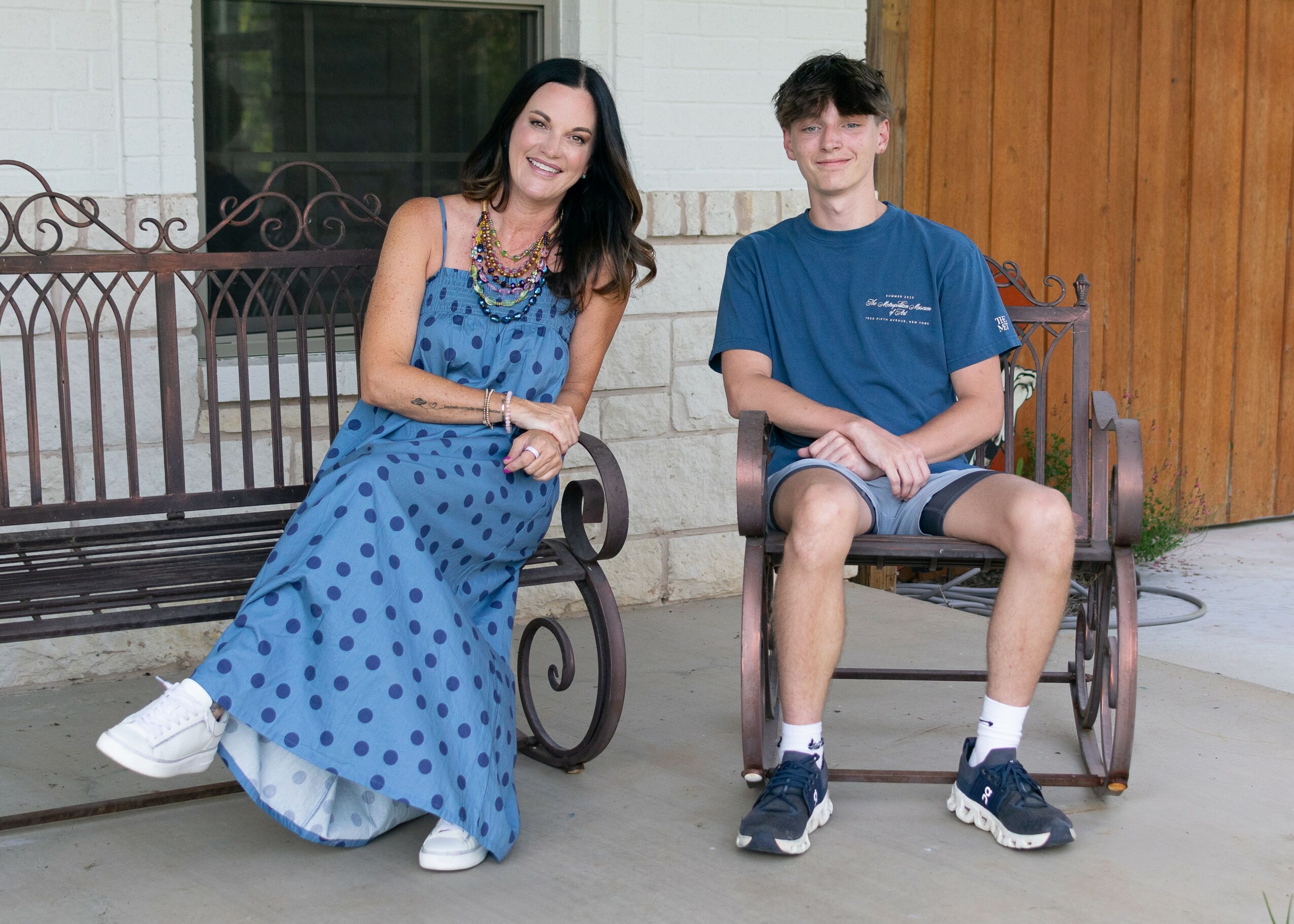 Woman and teenage boy sitting on benches outside, smiling, woman in a blue polka dot dress, boy in a blue T-shirt and shorts.