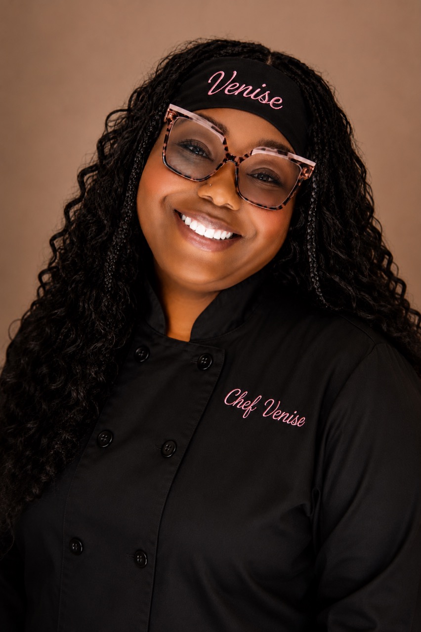 Smiling woman with curly hair, wearing glasses, a black headband with 'Venise', and a black chef's jacket with 'Chef Venise' embroidered.