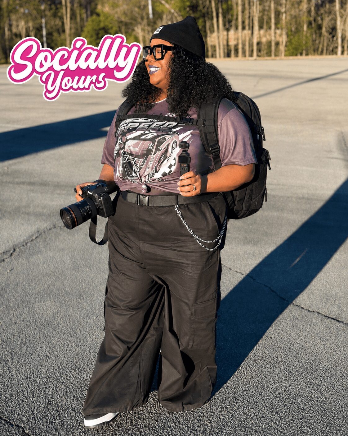 Woman with curly hair, glasses, black beanie, holding camera, standing outdoors with trees in background, smiling, carrying backpack.