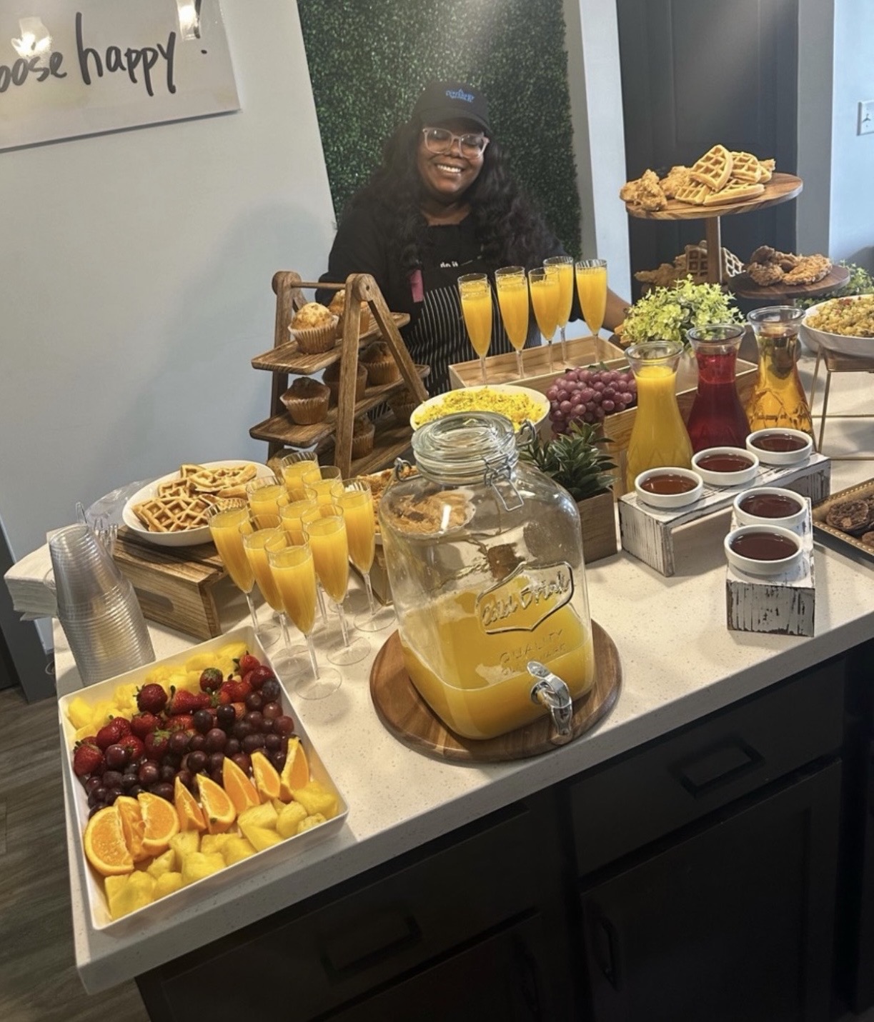 Woman smiling behind a breakfast buffet with orange juice, fruit, pastries, and condiments on a white counter.
