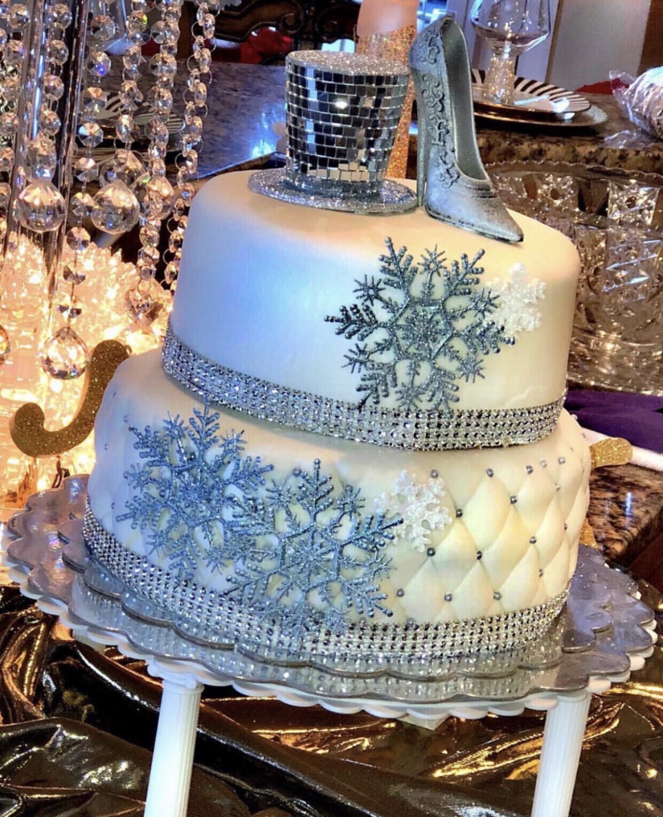 Two-tiered cake decorated with snowflakes, a silver shoe, and a glittery band, on a table with crystal decorations.