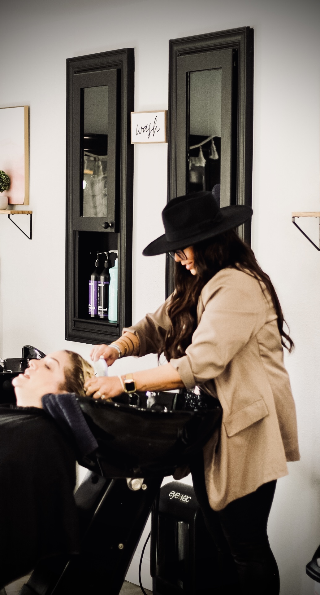 A person with long hair and a hat washes another person's hair at a salon sink, with mirrors and shelves in the background.