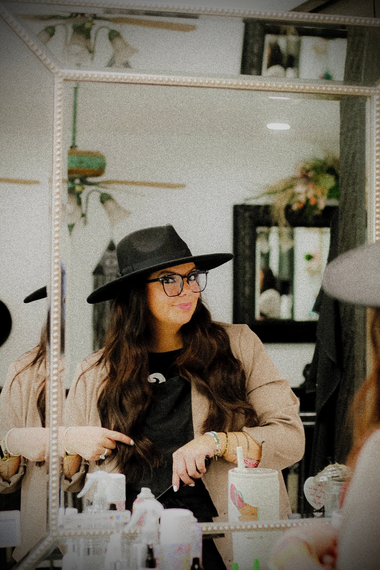 Woman with long hair, glasses, black hat, and beige jacket looking in mirror, surrounded by makeup items and decor.