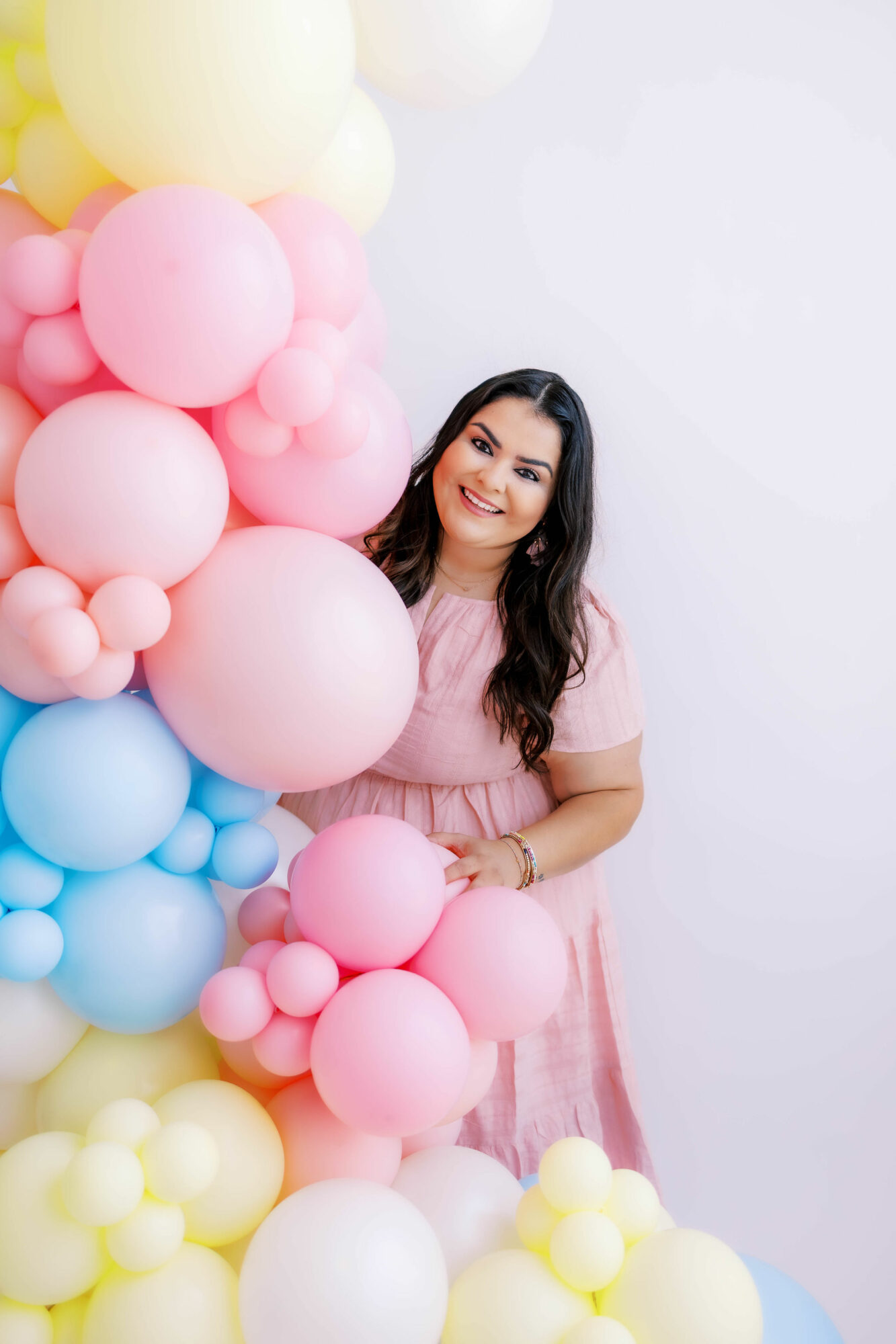 Smiling woman in pink dress standing next to colorful balloon arrangement against a white background.