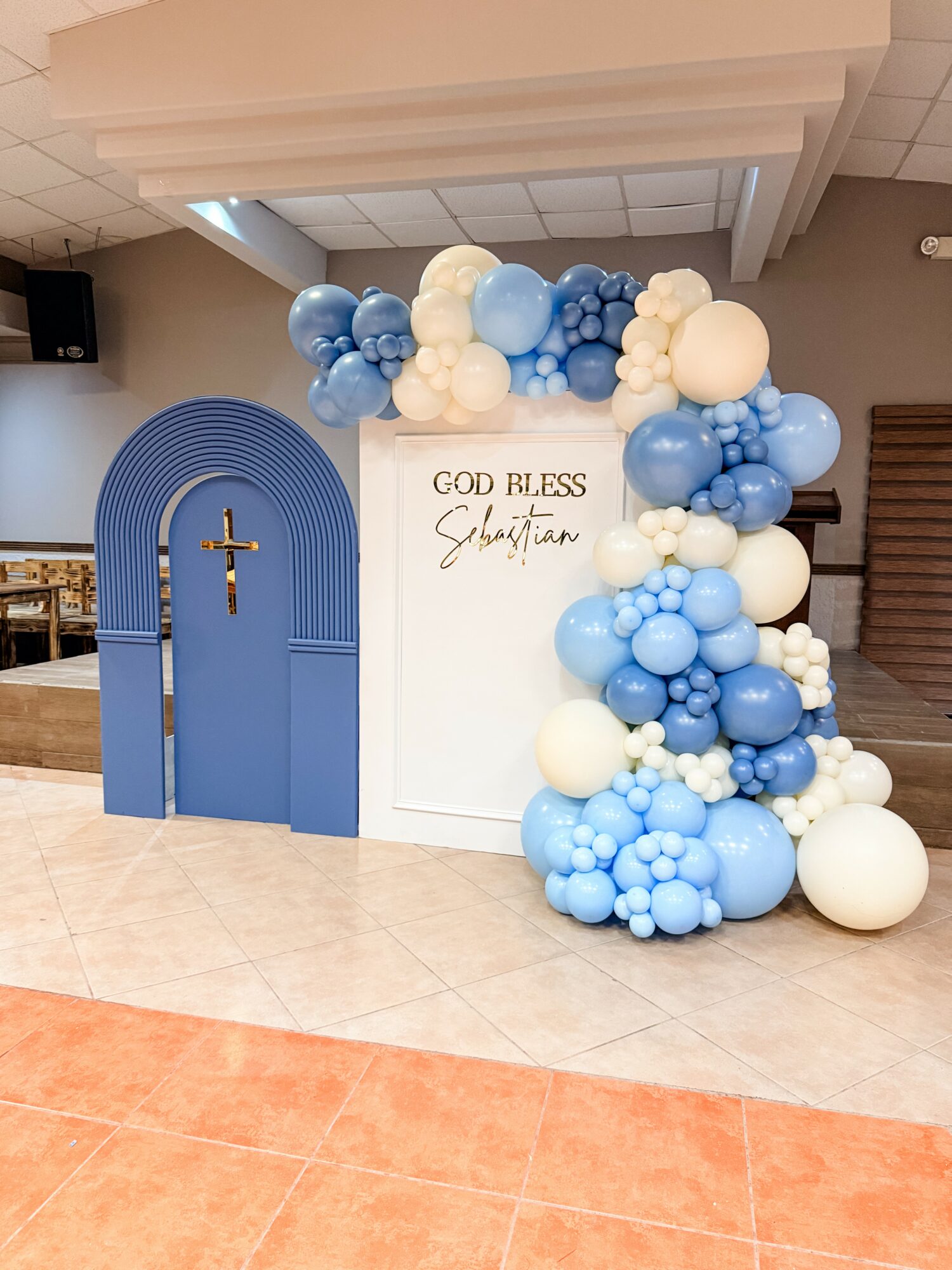 Decorative display with blue and white balloons, a white panel with gold text, and a blue arch with a cross, celebrating a religious event.