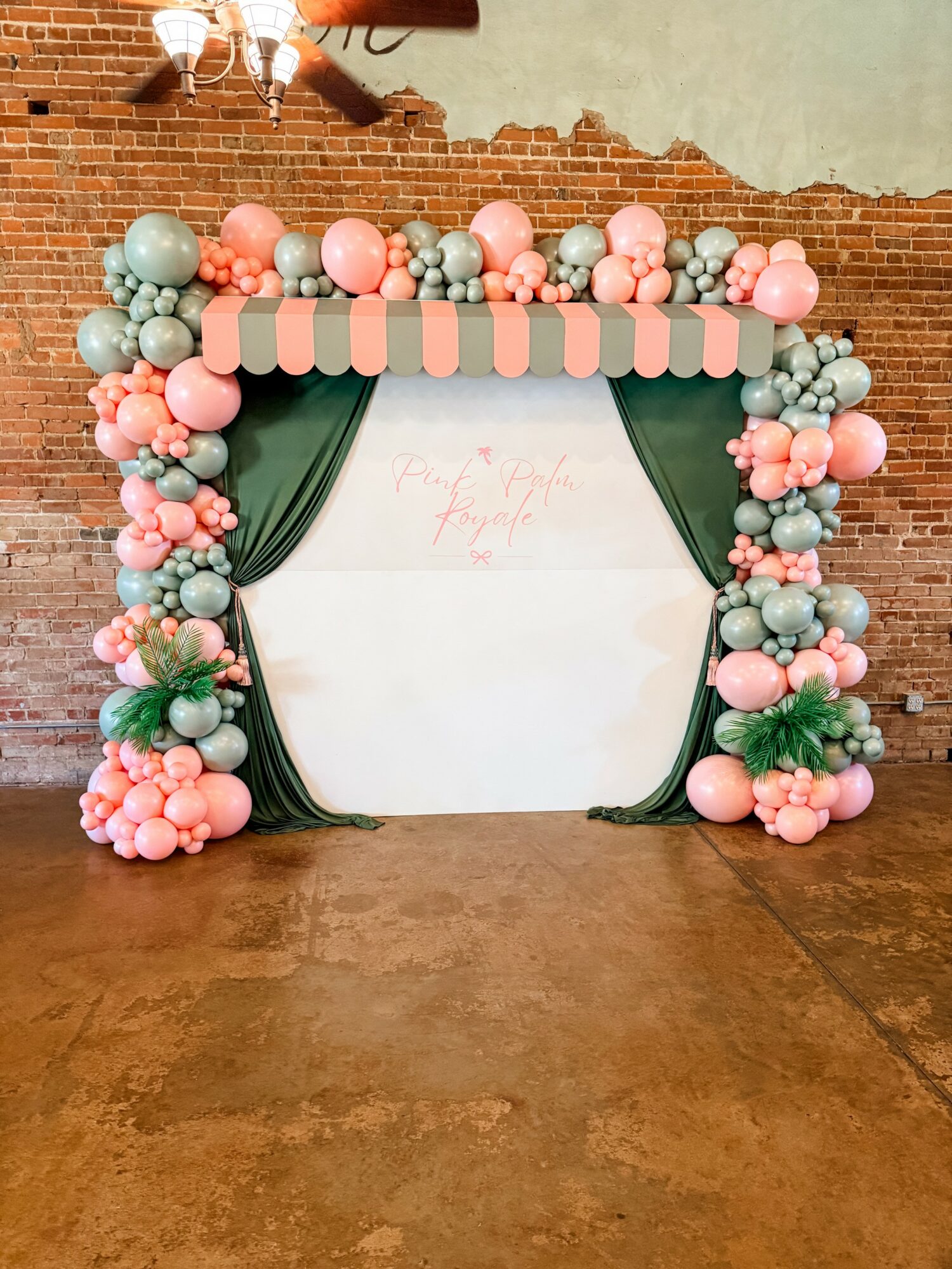 Balloon arch with pink, green, and peach balloons surrounds a white backdrop with green curtains, against a brick wall.