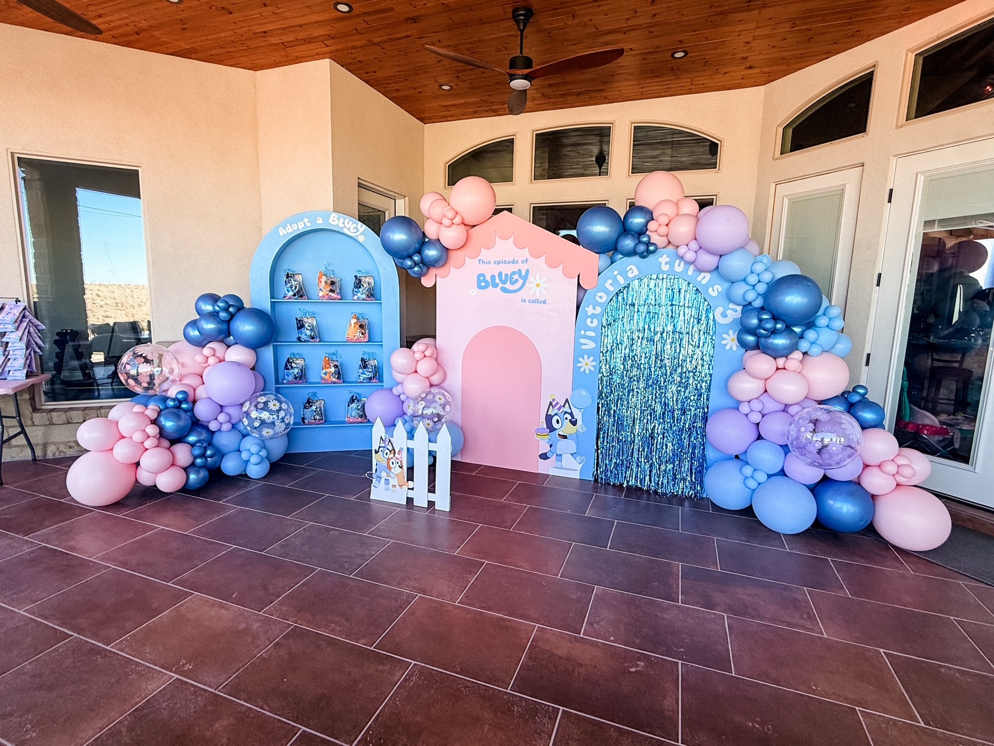 Decorative backdrop with balloons in pink, blue, and purple, featuring a pink arch, blue waterfall, and a small white fence, for a celebration.