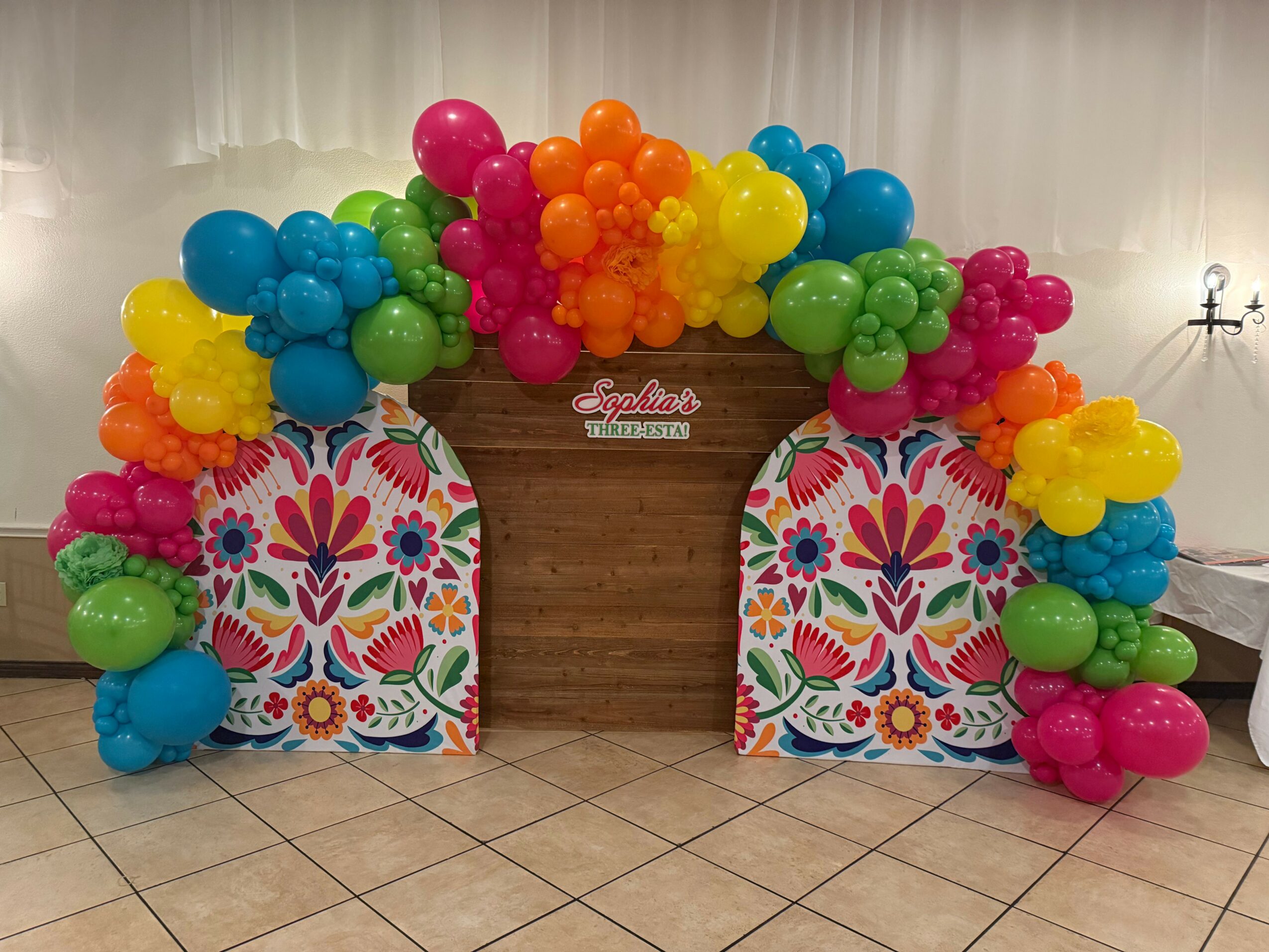 Colorful balloon arrangement with pink, orange, yellow, green, blue, and purple balloons around a wooden table with a floral patterned backdrop.