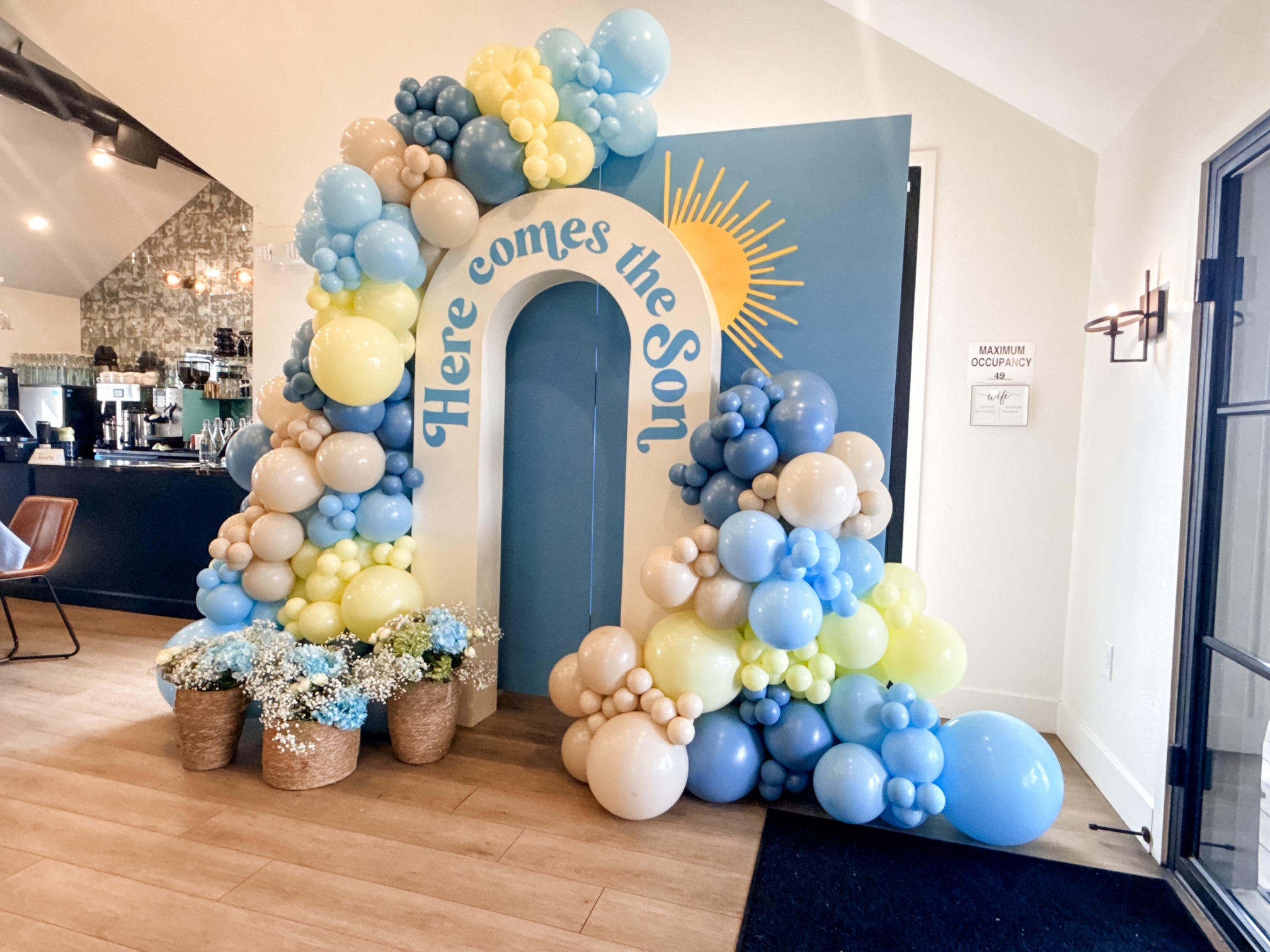 Decorative arch with blue, white, and yellow balloons, a large blue number one, and a sign reading 'Here comes the sun'.