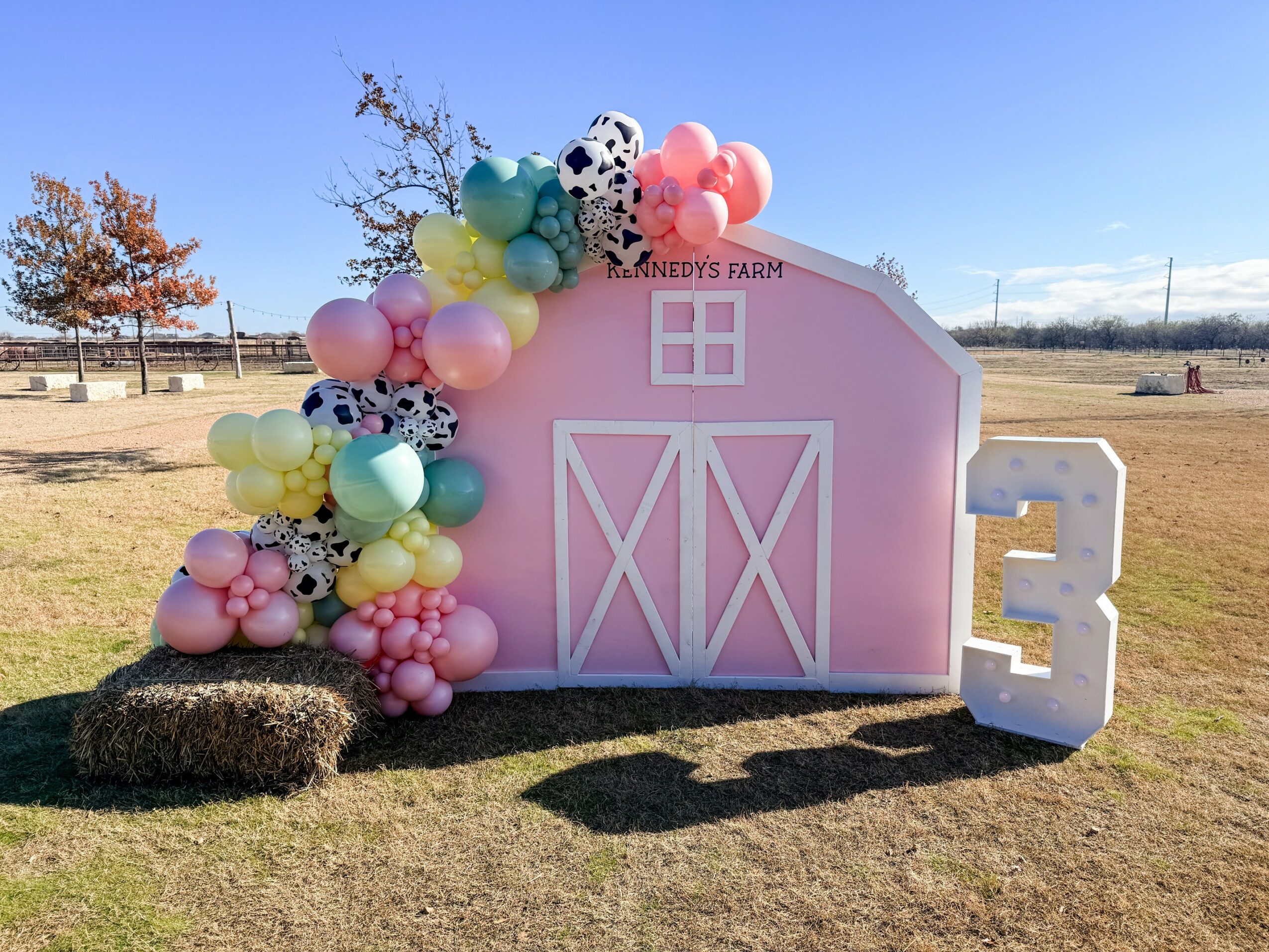 Pink barn-shaped decoration with balloons and a large number three on grass field, blue sky background.