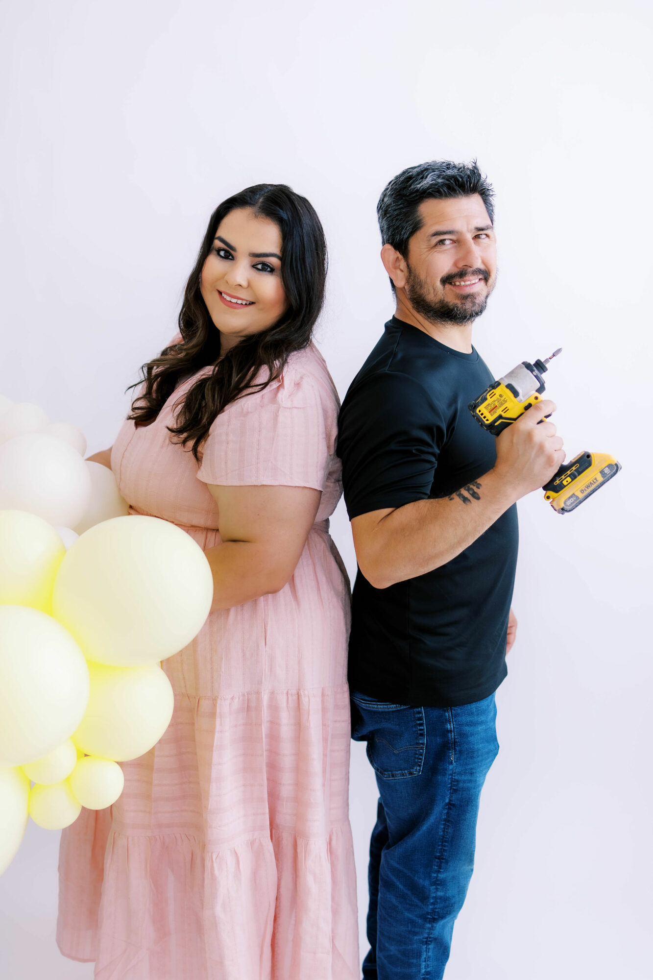 A woman in a pink dress and a man in a black shirt holding a drill, standing back-to-back, smiling, with balloons nearby.