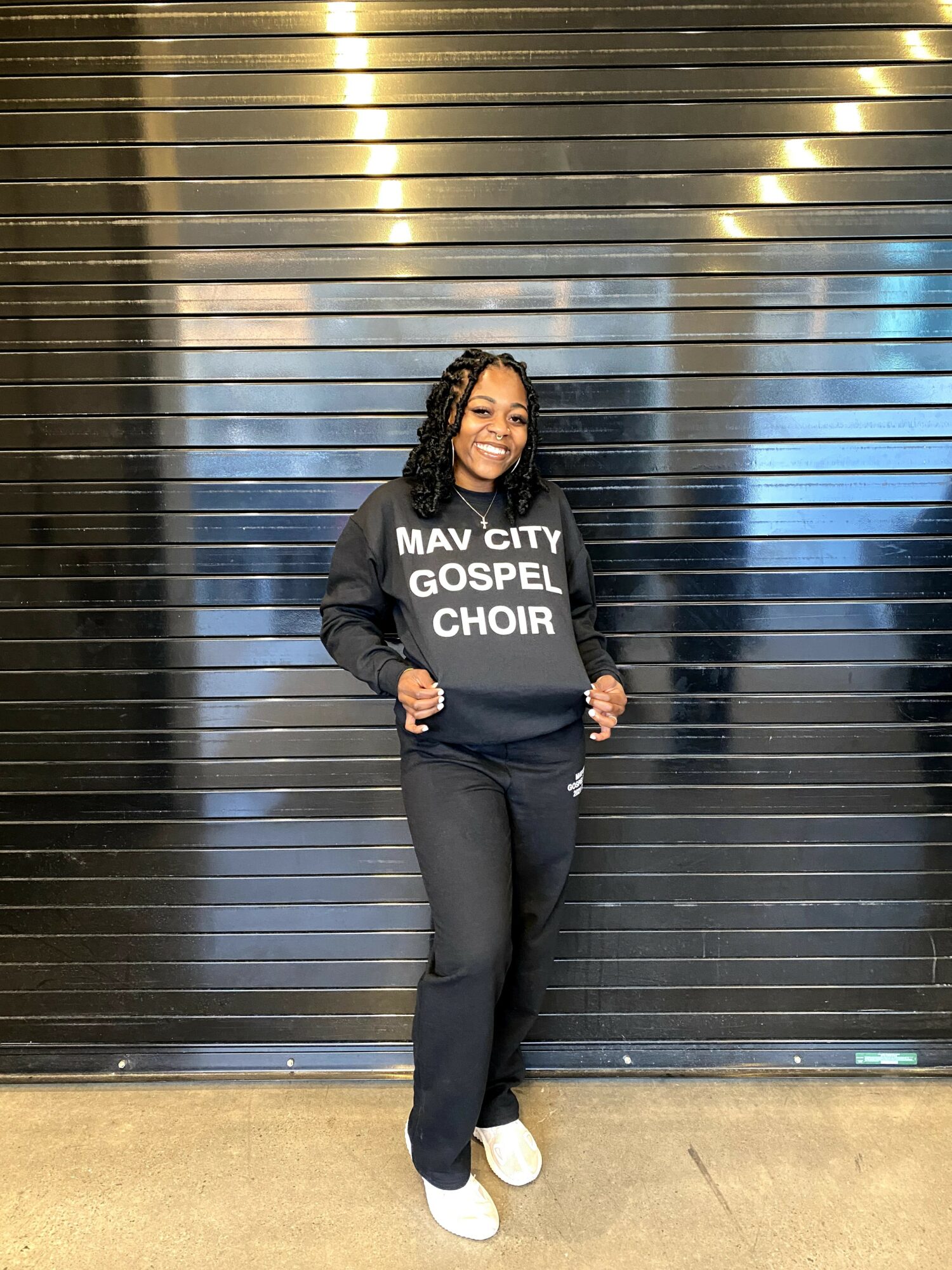 Woman smiling, wearing a black sweatshirt with white text, standing in front of a metallic, horizontal-striped background.