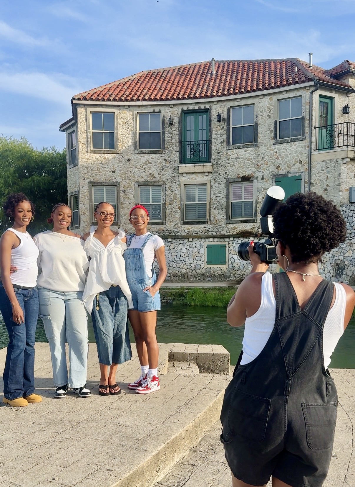Four women pose for a photo in front of a historic stone building with a red-tiled roof, beside a waterway.