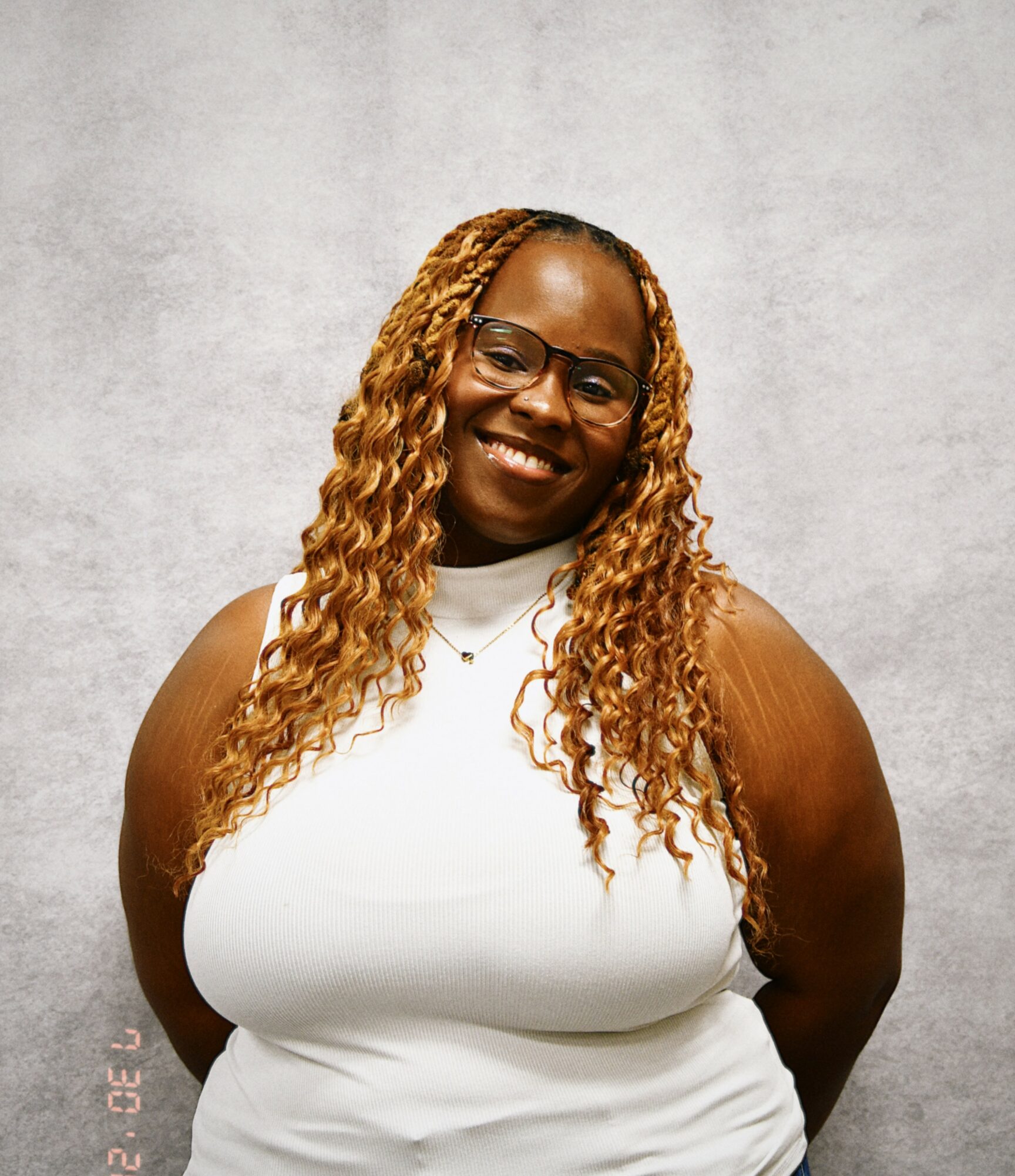 Smiling woman with glasses and long curly hair, wearing a white sleeveless top, standing against a light gray background.